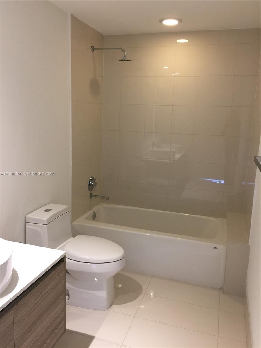 Full-height neutral wall tiling, bathtub with integrated shower, vanity with wood-finish cabinetry, white countertop, and recessed ceiling lighting