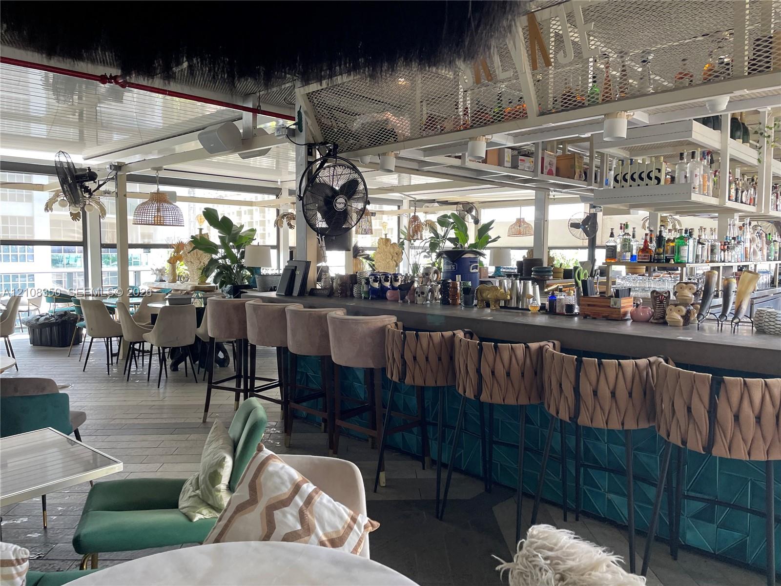Expansive bar area featuring a solid surface countertop, geometric teal tile facade, metal mesh overhead shelving, and industrial-style wall-mounted fans