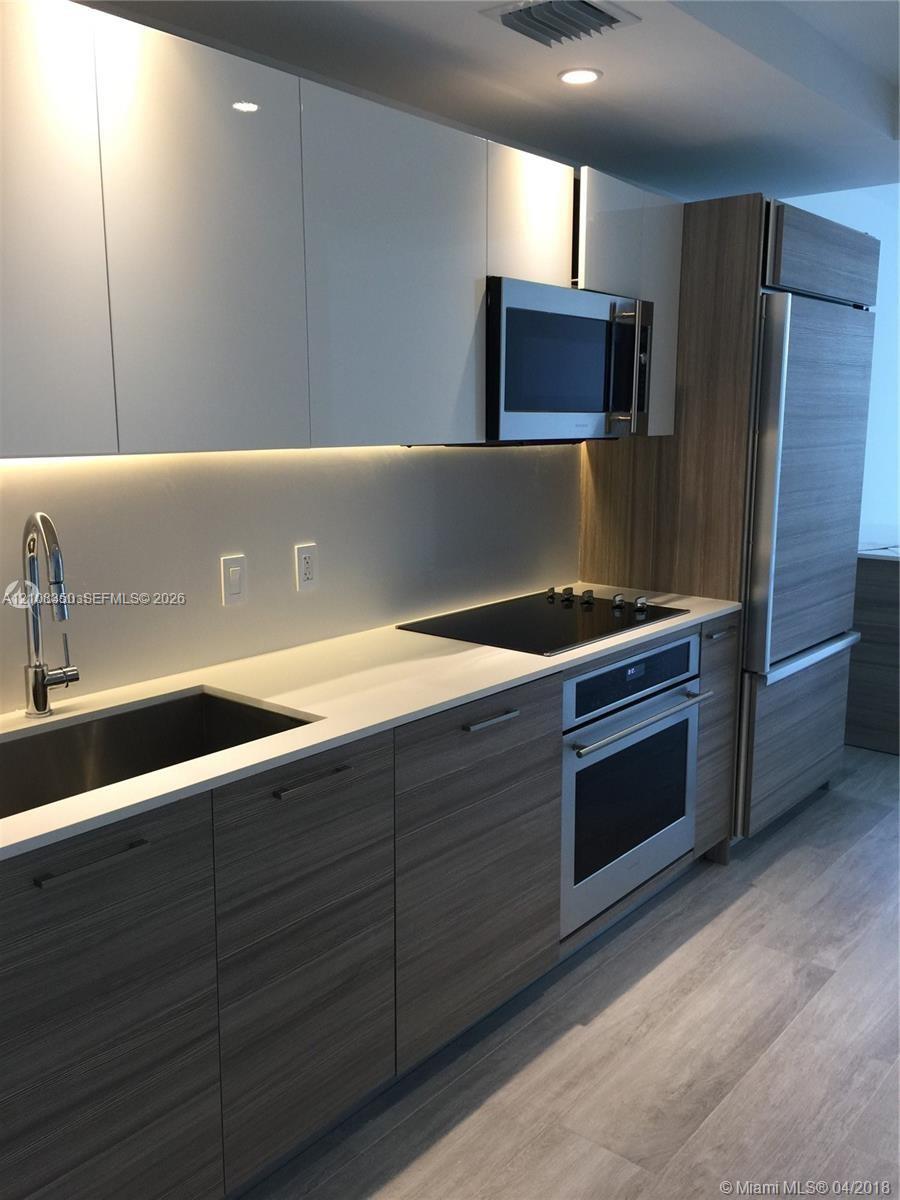 Kitchen featuring wood-finish lower cabinetry, white gloss upper cabinetry, stone countertops, stainless steel appliances, and integrated under-cabinet lighting