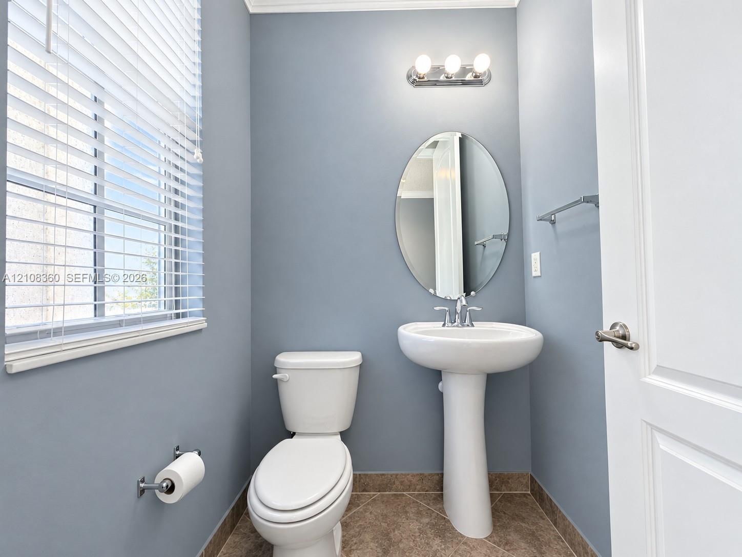 White pedestal sink with chrome faucet, oval wall-mounted mirror, three-light vanity fixture, and tile flooring
