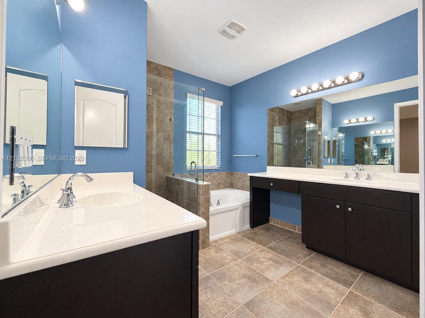 Bathroom featuring a dual-sink vanity with dark cabinetry, solid-surface countertops, large wall-mounted mirrors, a tiled walk-in shower with glass enclosure, and a soaking tub