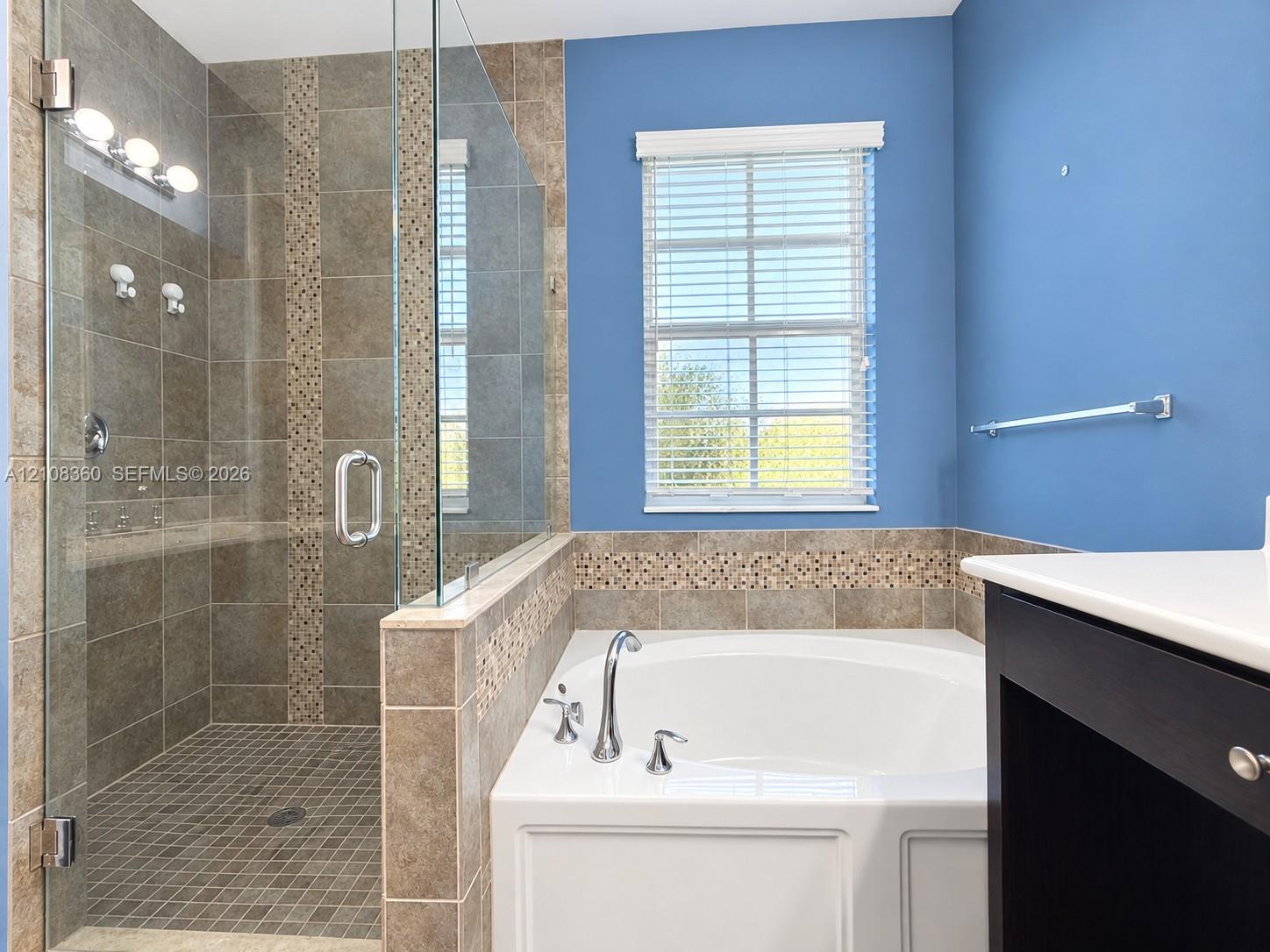 Walk-in shower with glass enclosure, soaking tub with deck-mounted faucet, tile surround with mosaic accent detailing, and dark-finish vanity