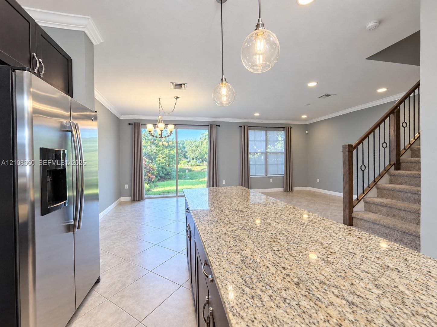 Kitchen featuring a sprawling stone-surface island, dark-toned cabinetry, stainless steel refrigerator, glass pendant lighting, and neutral floor tile Kitchen featuring a sprawling stone-surface island, dark-toned cabinetry, stainless steel refrigerator, glass pendant lighting, and neutral floor tile