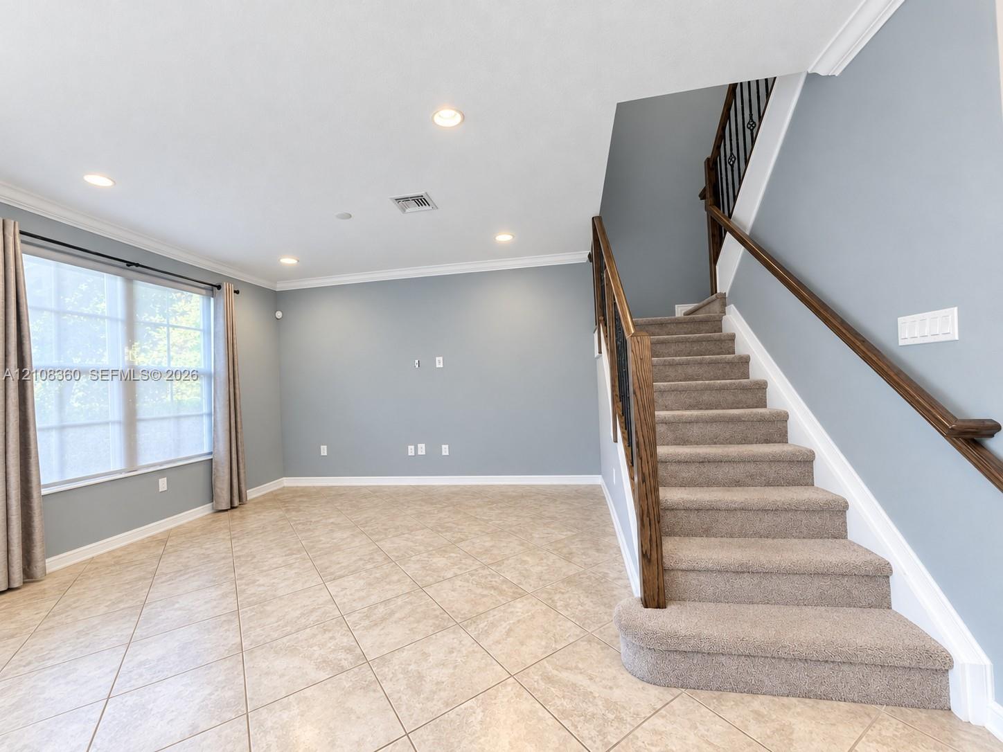 Open interior space featuring neutral tile flooring, recessed lighting, crown molding, and a staircase with a wooden handrail and metal balusters Open interior space featuring neutral tile flooring, recessed lighting, crown molding, and a staircase with a wooden handrail and metal balusters