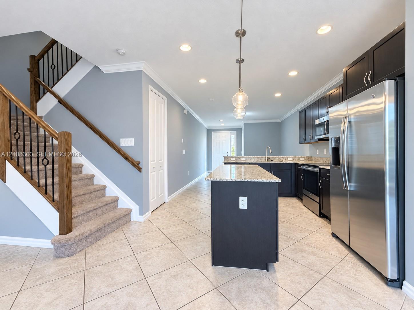 Kitchen featuring dark cabinetry, stone-finish countertops, stainless steel appliances, and a center island, complemented by a staircase with wooden handrails and iron balusters Kitchen featuring dark cabinetry, stone-finish countertops, stainless steel appliances, and a center island, complemented by a staircase with wooden handrails and iron balusters