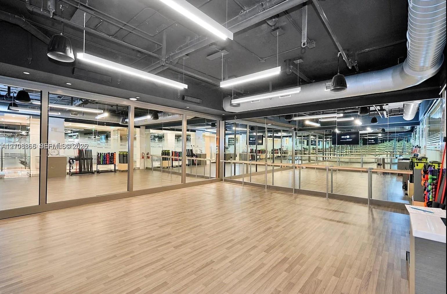 Studio space featuring wood-finish flooring, floor-to-ceiling mirrors, exposed industrial ceiling, linear LED lighting, and a wall-mounted wooden ballet barre