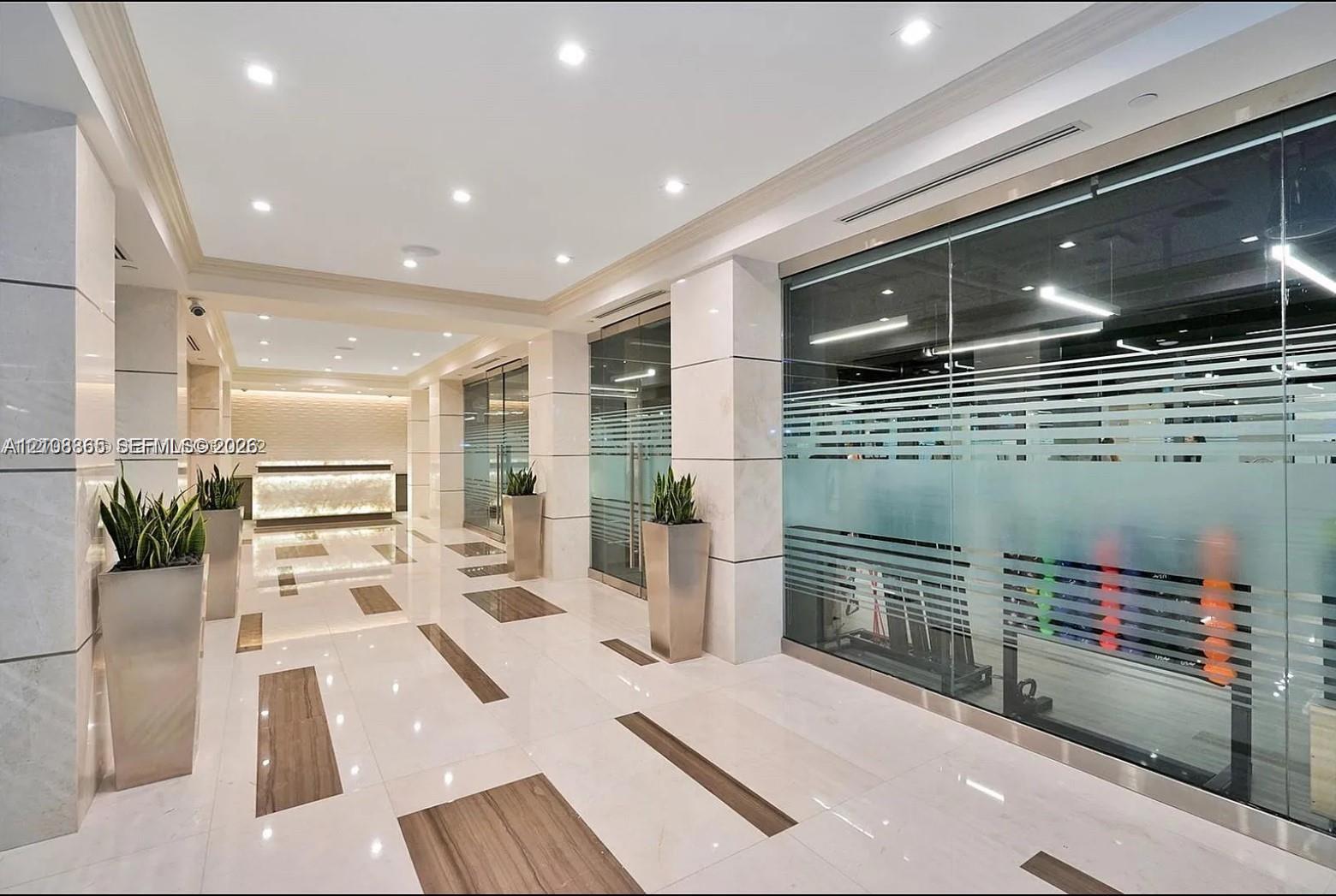 Polished stone flooring with inlaid wood-finish accents, large-format stone pillar cladding, glass-partitioned wall panels, recessed lighting, and a backlit stone-front desk