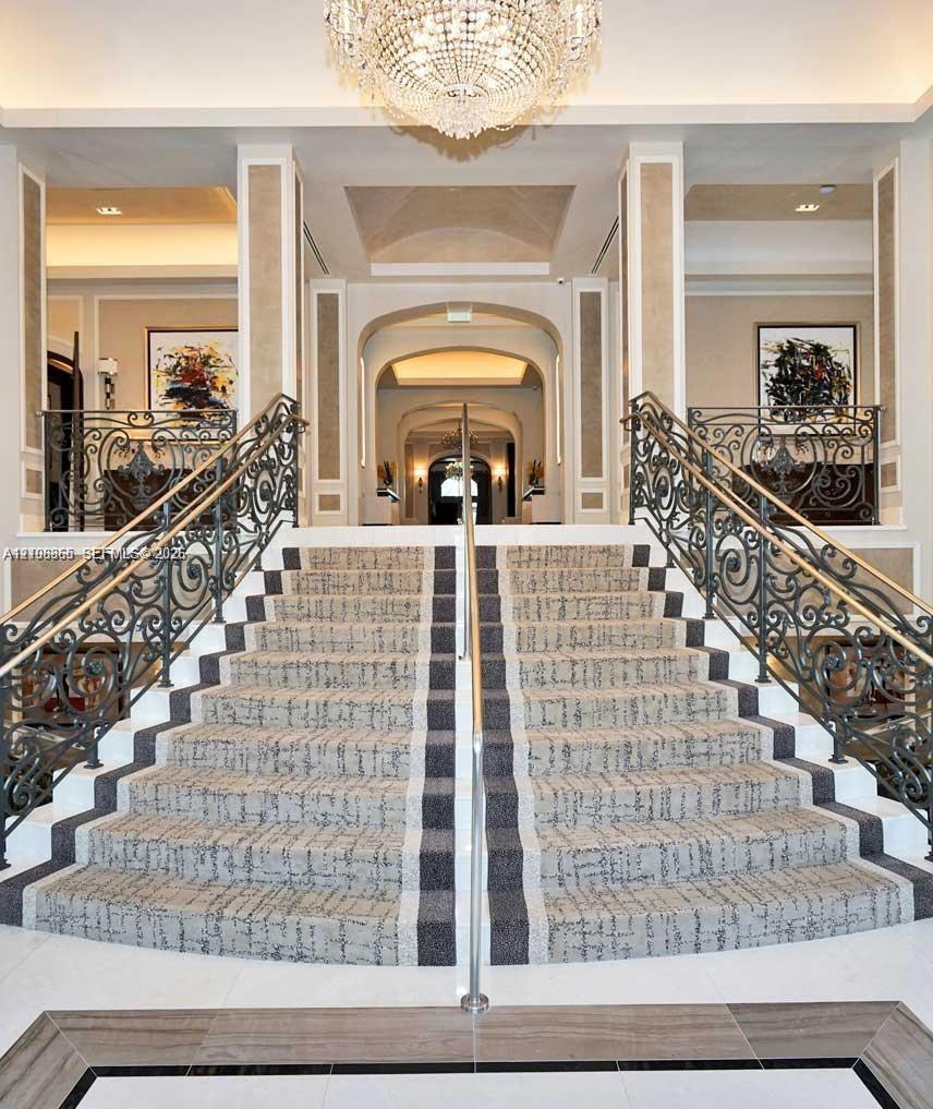 Grand dual staircase featuring intricate wrought iron railings, stone flooring with decorative inlay, and a central crystal chandelier under vaulted ceiling detailing