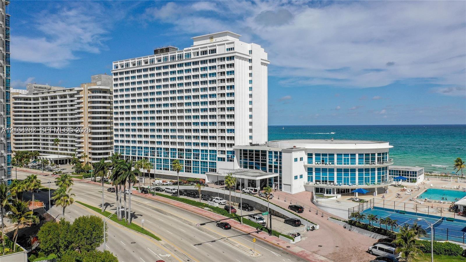 High-rise residential exterior featuring expansive glass facade, swimming pool, and tennis court, situated along the coastline