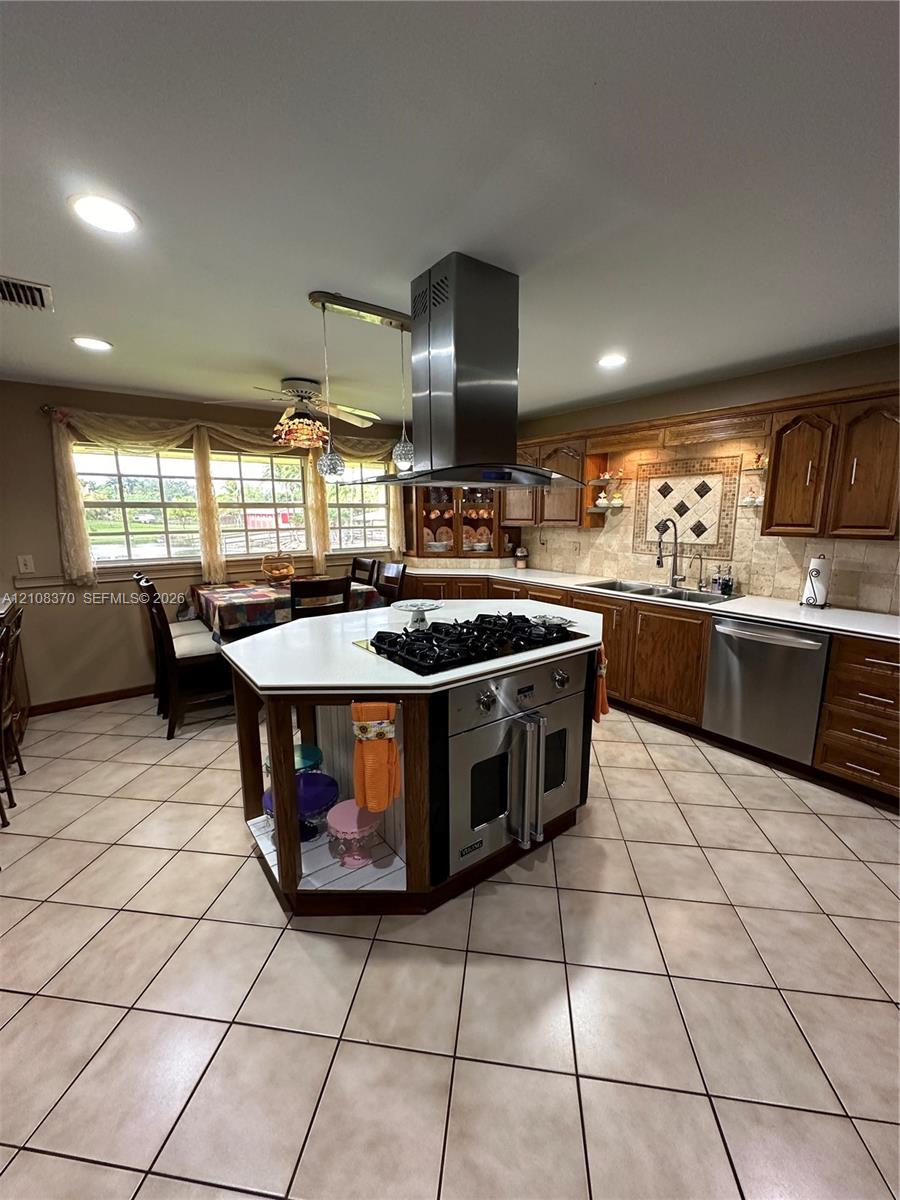 Kitchen featuring a central island with a built-in gas cooktop, stainless steel ventilation hood, wood cabinetry, stone-finish tile flooring, and recessed lighting