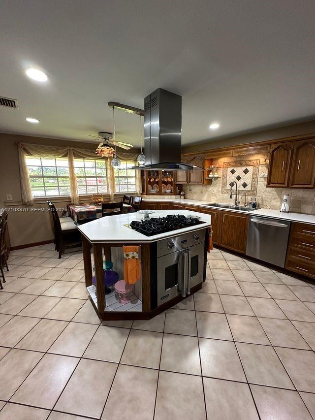 Kitchen featuring a hexagonal island with an integrated cooktop and oven, stainless steel range hood, stone-finish tile flooring, recessed lighting, and wood cabinetry
