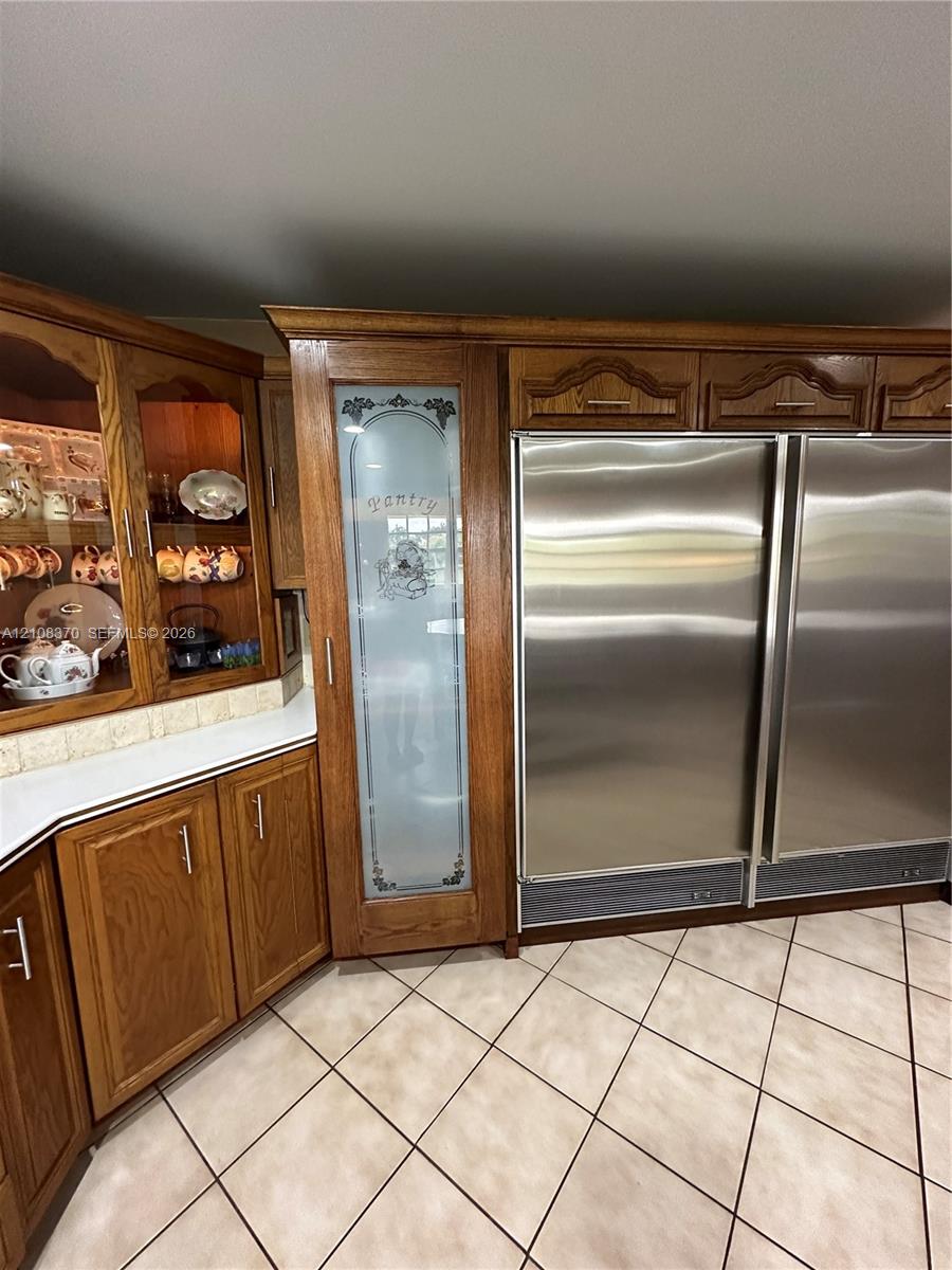 Custom wood cabinetry, floor-to-ceiling pantry with etched glass door, integrated side-by-side stainless steel refrigeration, and ceramic tile flooring