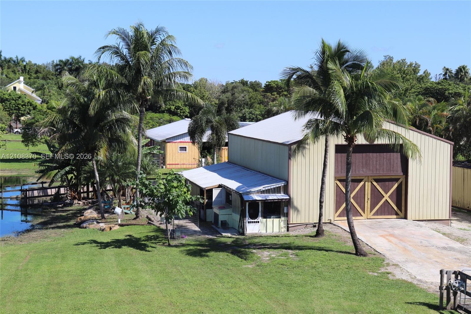Metal-sided workshop featuring large carriage-style doors, a concrete driveway, and mature palm tree landscaping on a waterfront lot