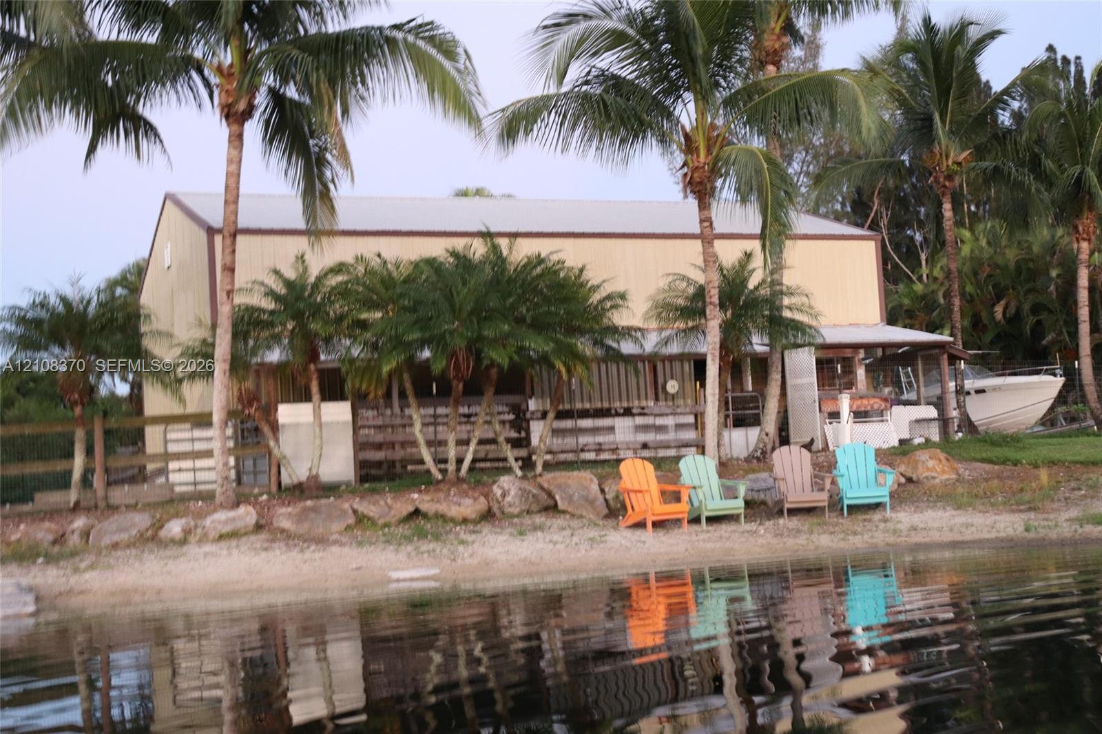 Waterfront structure featuring a metal roof, stacked stone riprap, and perimeter fencing surrounded by mature palm trees