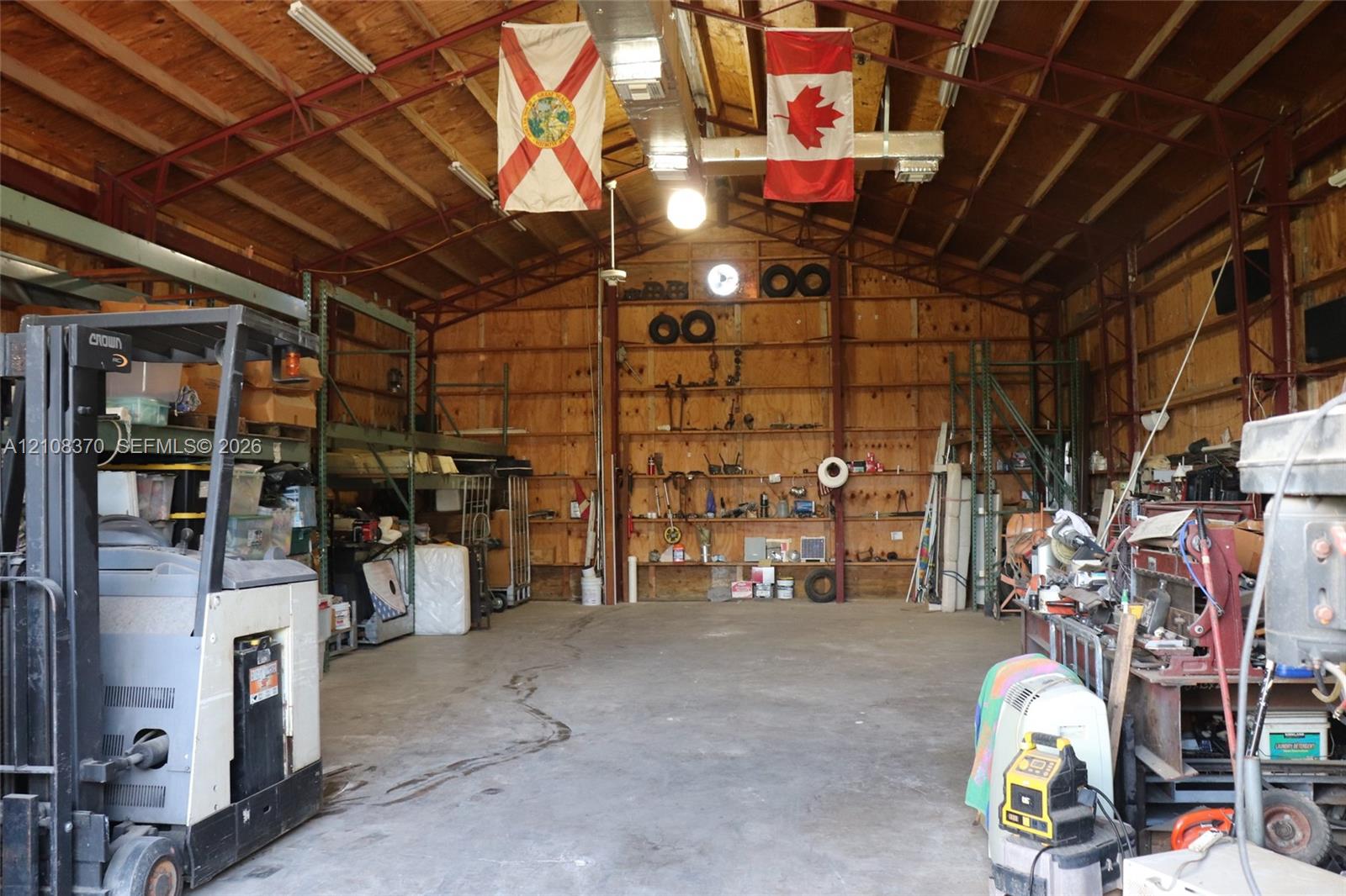 Expansive metal-framed workshop featuring exposed truss construction, high ceilings, concrete flooring, plywood wall paneling, and integrated fluorescent lighting