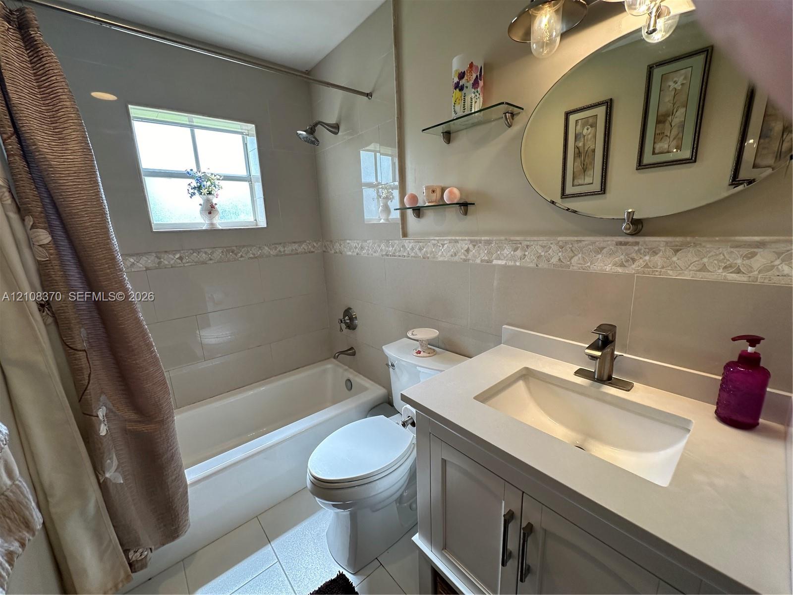 Bathroom featuring a vanity with a solid-surface countertop, an integrated sink, an oval wall-mounted mirror, neutral tile wall surround with decorative accent inlay, and a tub-shower combination