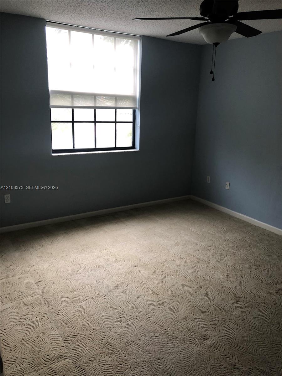 Room featuring patterned carpeting, a multi-pane window with a roller shade, textured ceiling, and a ceiling fan with integrated light fixture