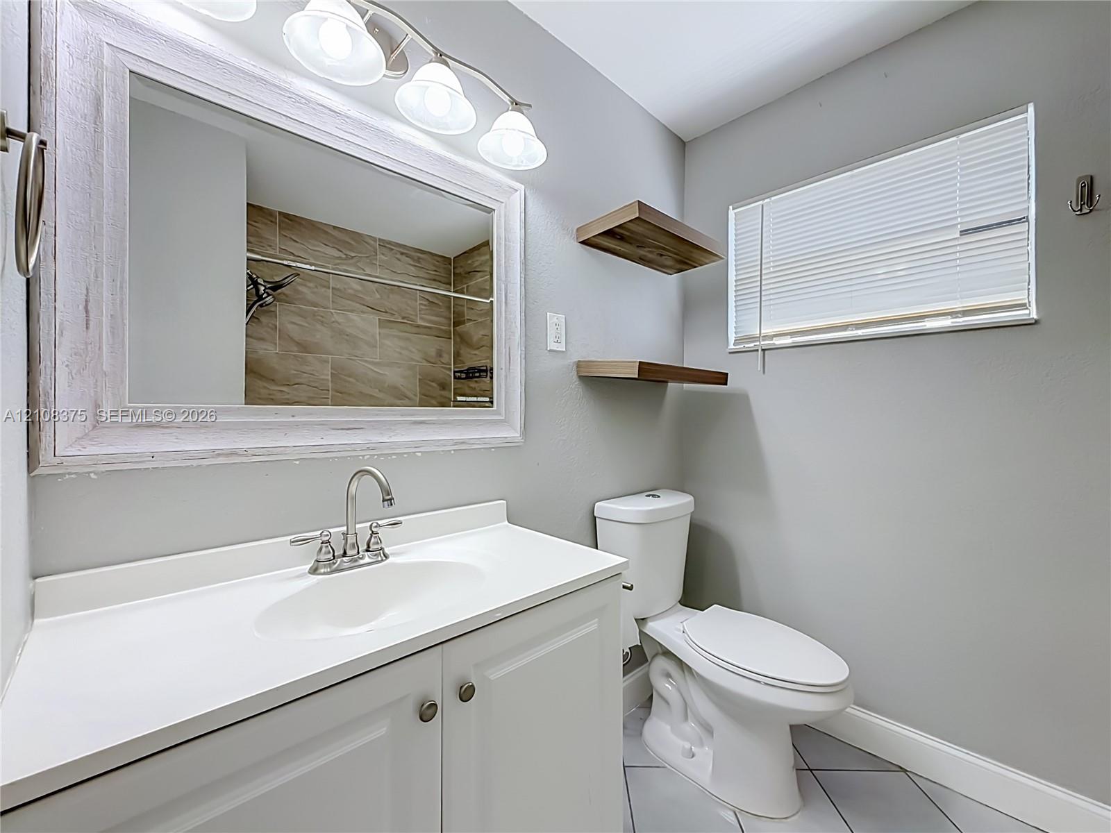 Bathroom featuring a white vanity cabinet with a solid-surface countertop, an integrated sink, a framed mirror with a four-light fixture, wood-finish floating shelves, and tile flooring