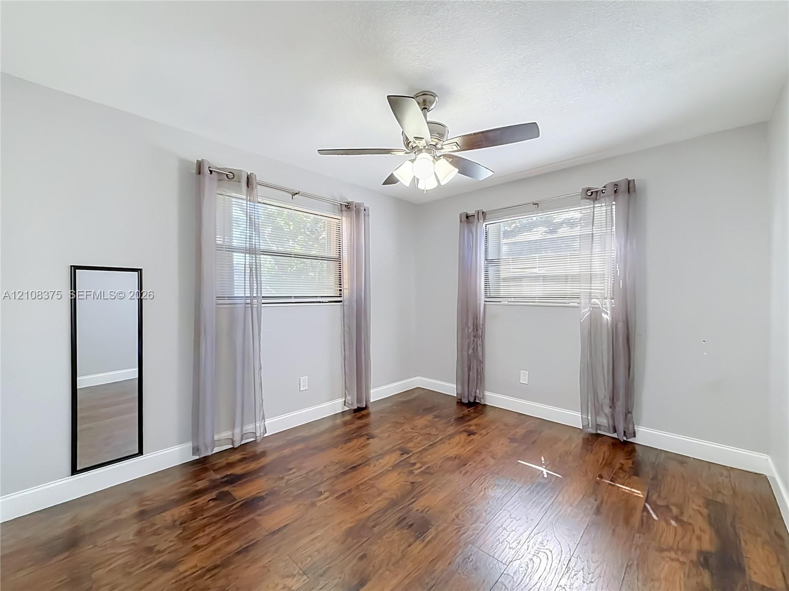 Wood-finish flooring, dual windows with horizontal blinds, and a central ceiling fan with integrated lighting