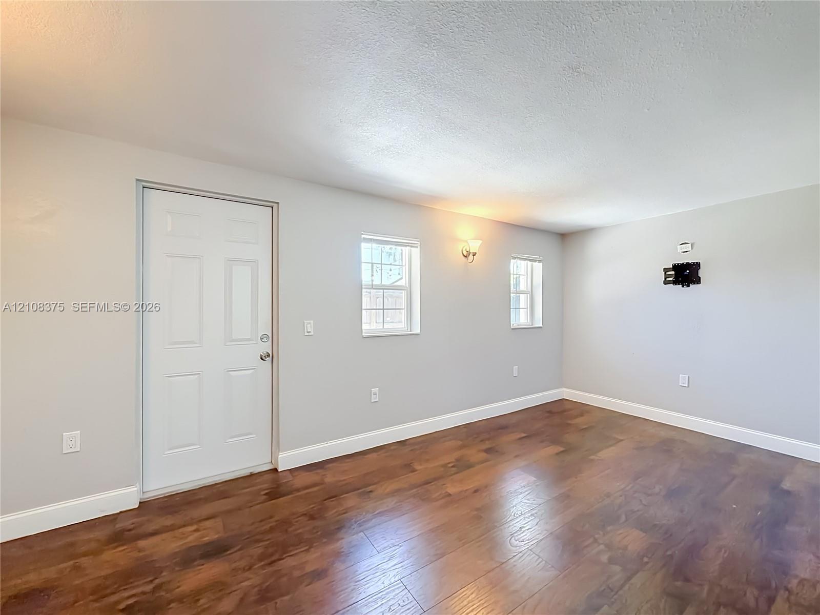 Interior space featuring wood-finish flooring, neutral wall paint, two double-hung windows, a solid-core entry door, and wall-mounted lighting