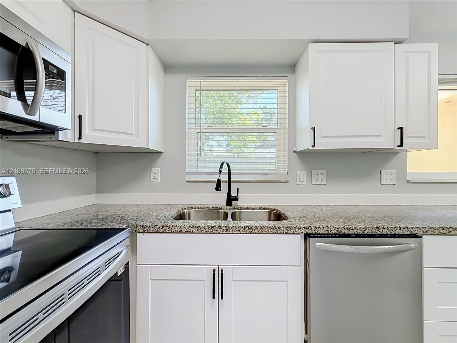 Kitchen featuring white shaker-style cabinetry, stone-finish countertops, stainless steel appliances, dual-basin stainless steel sink with matte black gooseneck faucet, and a window with blinds