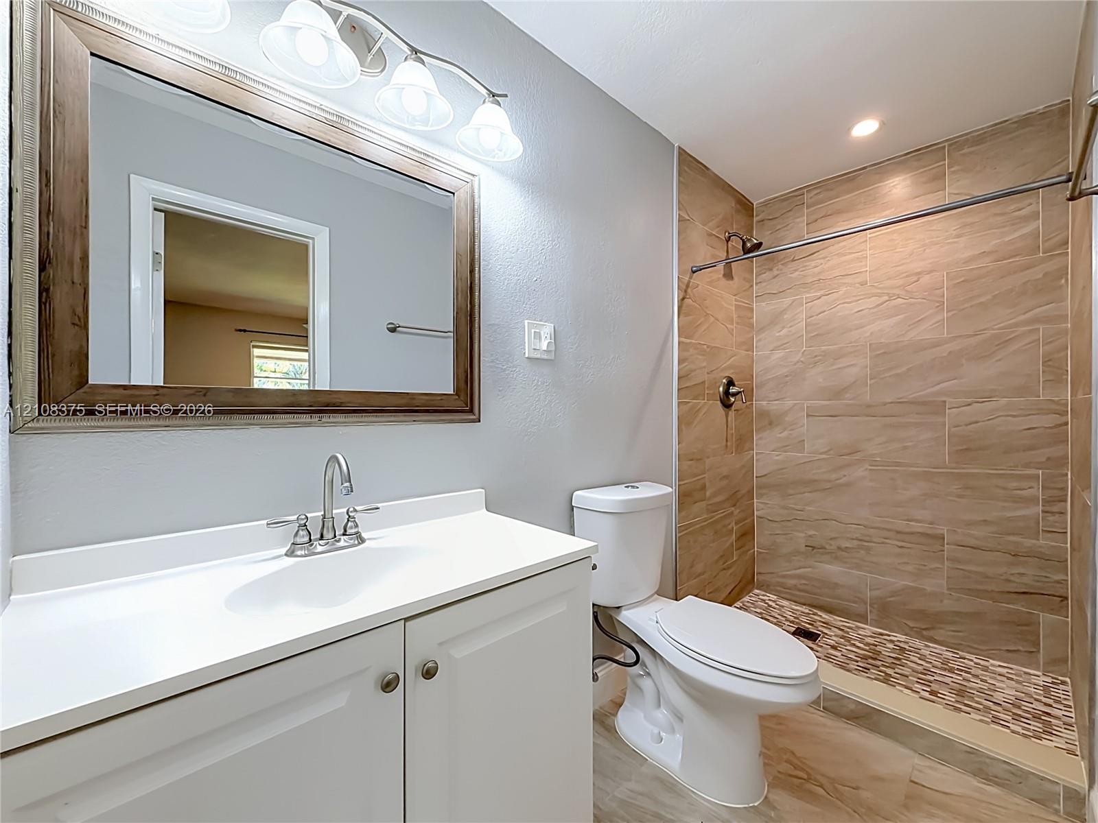 Bathroom featuring a vanity with a white countertop and sink, walk-in shower with horizontal tile surround, mosaic stone-finish shower floor, and a three-light wall-mounted fixture