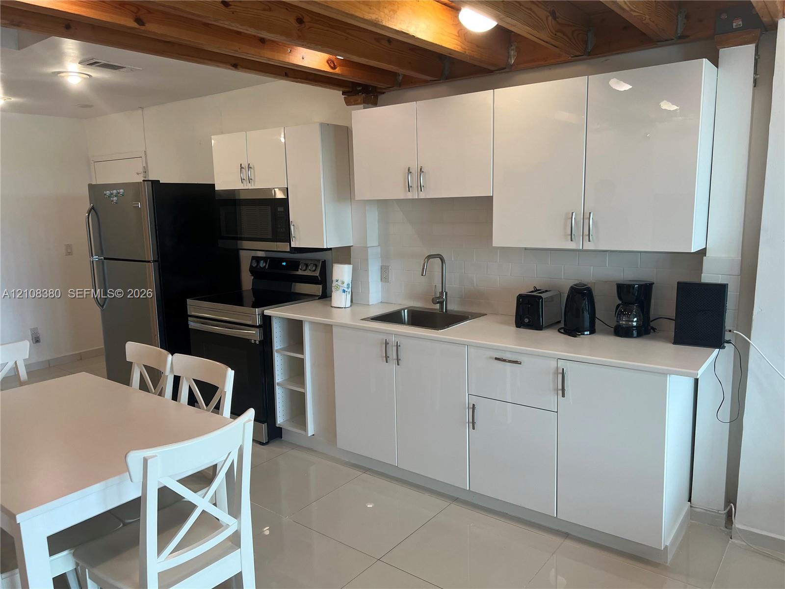 Kitchen featuring high-gloss white cabinetry, white stone-finish countertops, exposed wooden ceiling beams, and polished tile flooring Kitchen featuring high-gloss white cabinetry, white stone-finish countertops, exposed wooden ceiling beams, and polished tile flooring