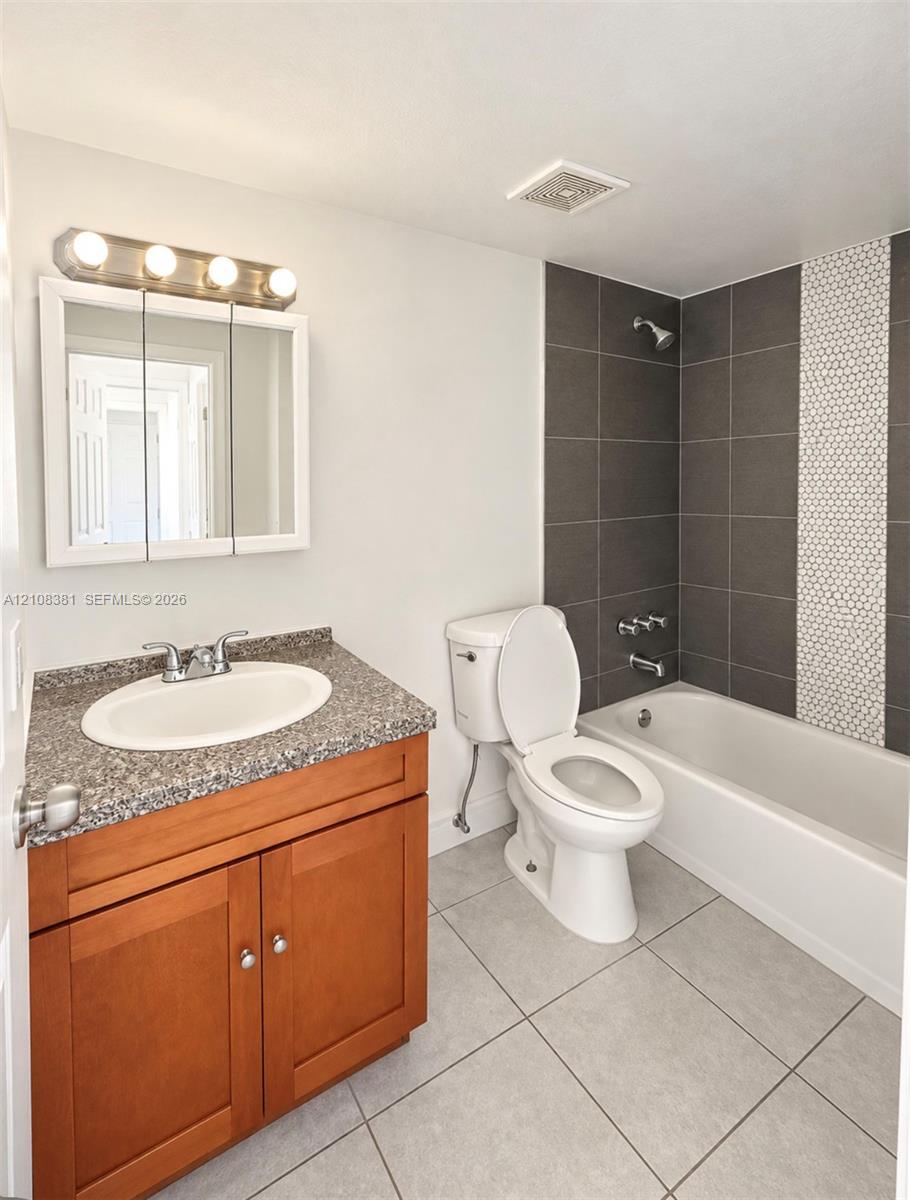 Bathroom featuring a wood-finish vanity with a stone-composite countertop, integrated medicine cabinet, tile-surround tub with a decorative mosaic accent strip, and large-format floor tiling