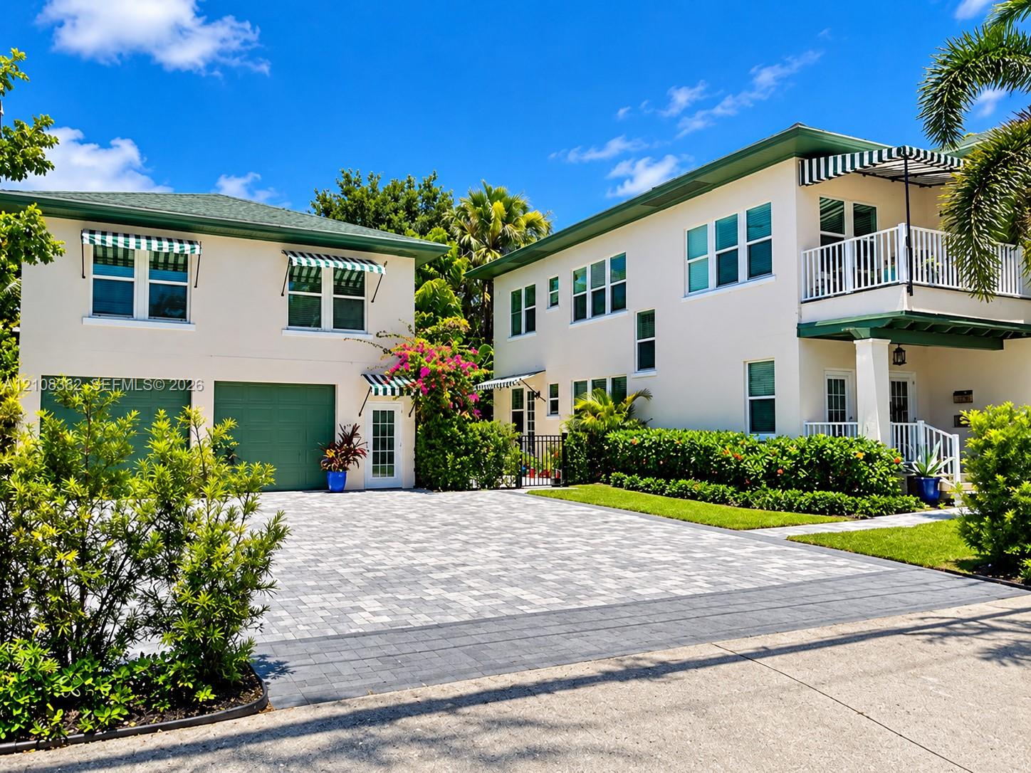 Two-story exterior featuring multi-tone paver driveway, green-tinted garage doors, striped fabric awnings, a second-story balcony with white railing, and structured landscaping Two-story exterior featuring multi-tone paver driveway, green-tinted garage doors, striped fabric awnings, a second-story balcony with white railing, and structured landscaping