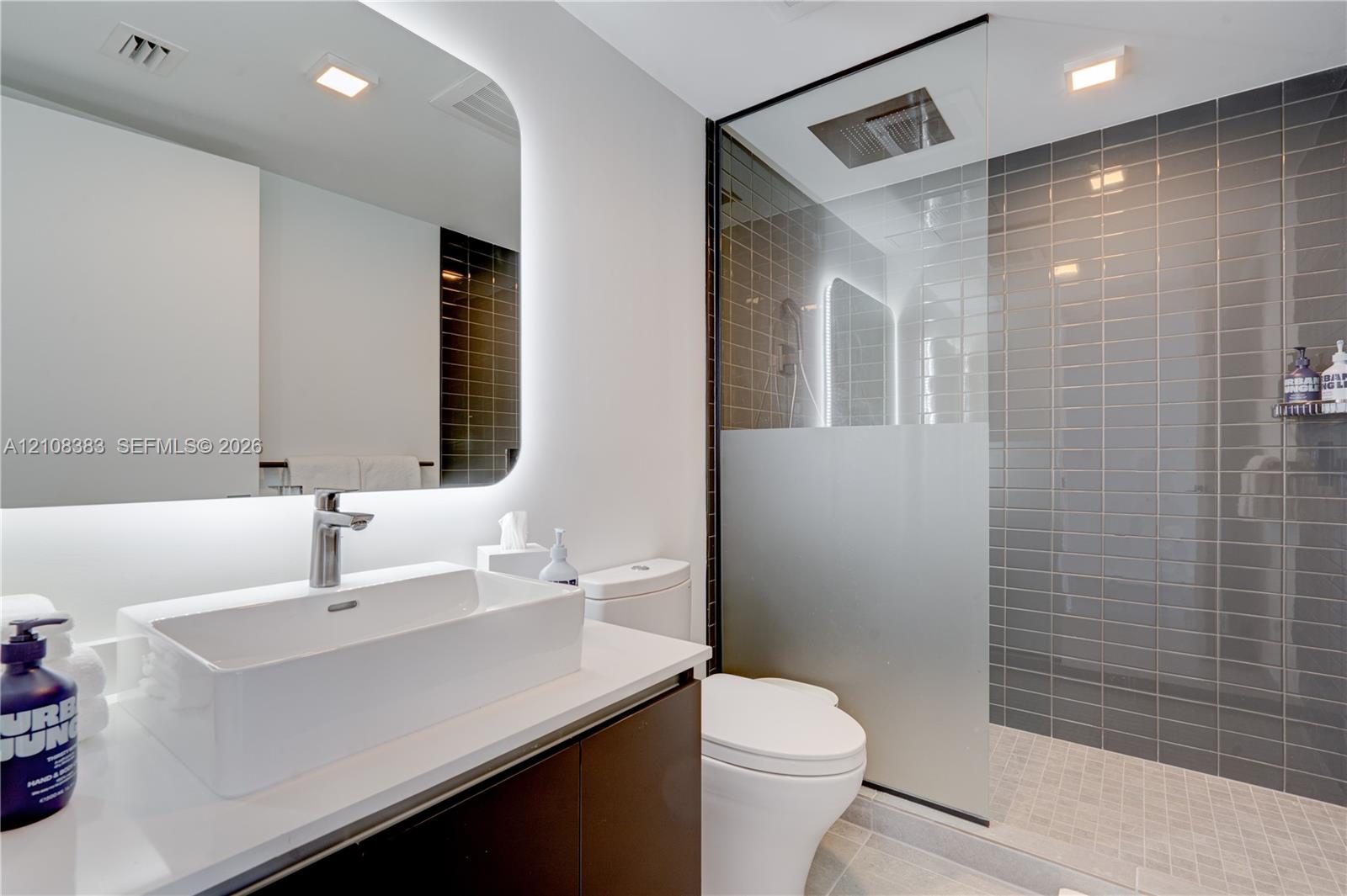 Modern vanity with rectangular vessel sink, polished countertop, and backlit mirror, alongside a walk-in shower featuring a glass enclosure with frosted paneling and gray subway tile surround Modern vanity with rectangular vessel sink, polished countertop, and backlit mirror, alongside a walk-in shower featuring a glass enclosure with frosted paneling and gray subway tile surround