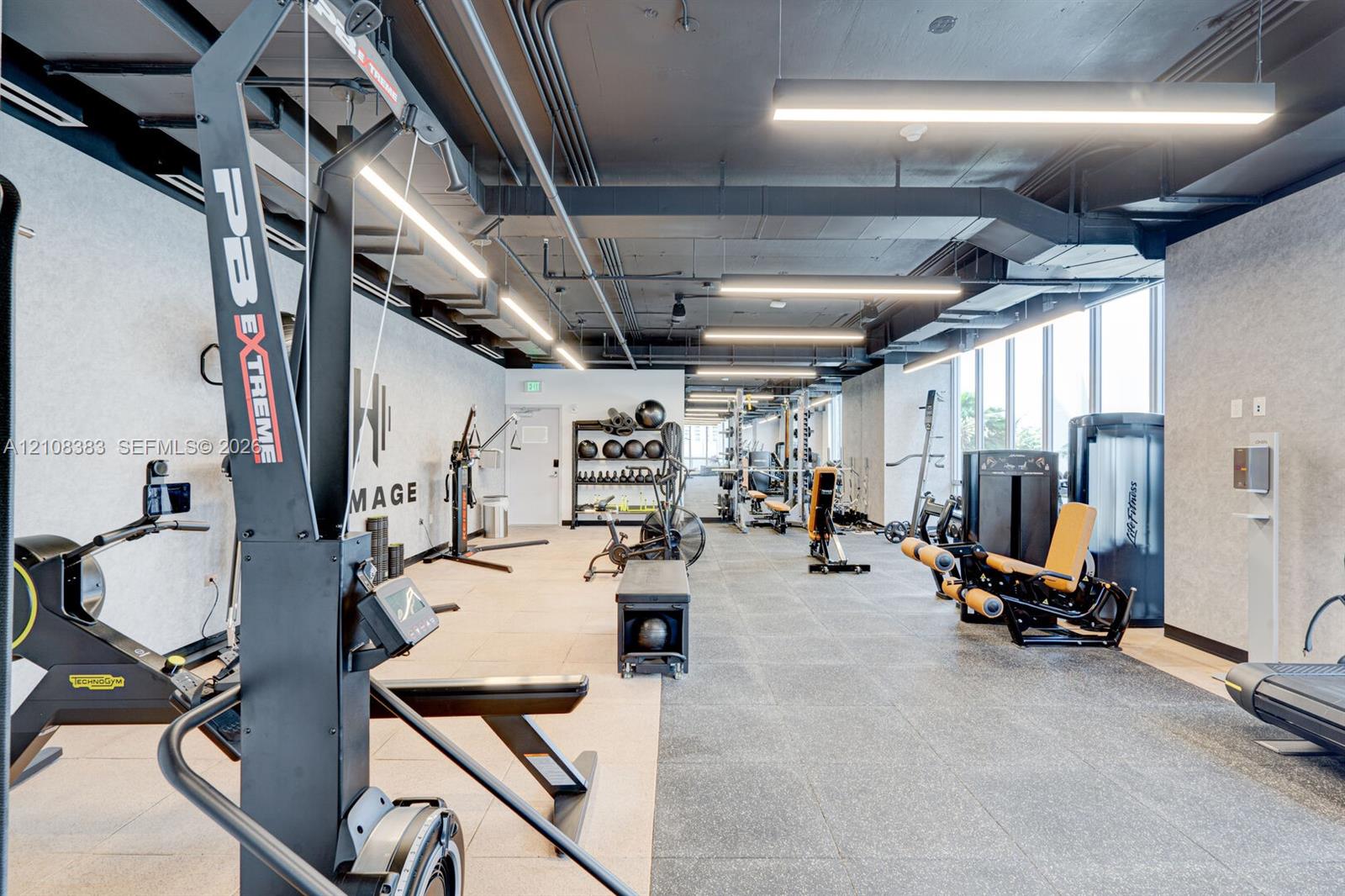Fitness center with industrial-style exposed ceiling, recessed linear lighting, rubberized flooring, and expansive floor-to-ceiling windows