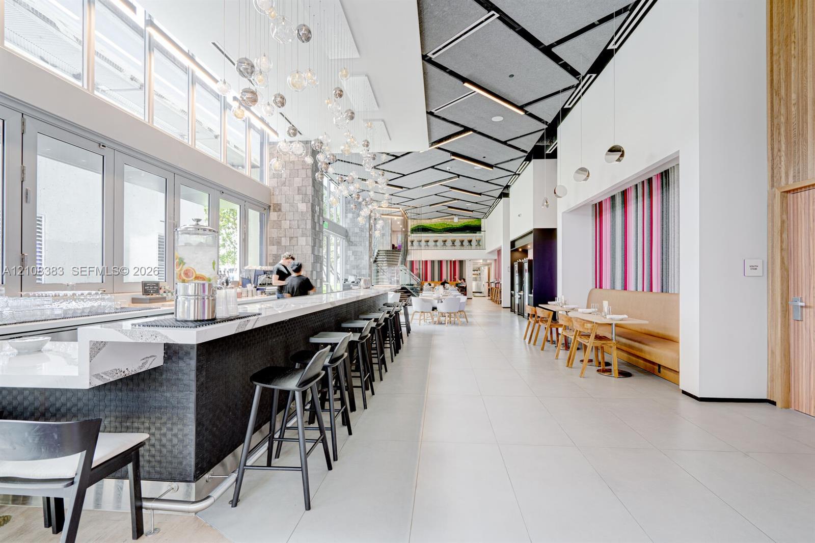 Large-scale bar area featuring a polished countertop, metallic front paneling, high-ceiling geometric acoustic panels, floor-to-ceiling glass windows, and custom multi-pendant lighting