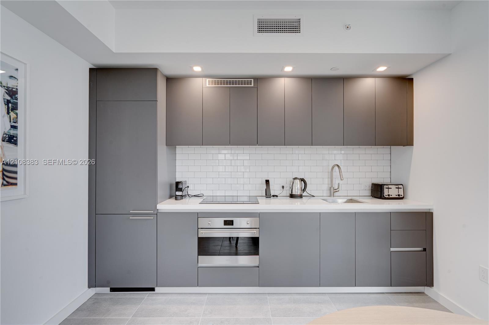 Kitchen featuring flat-panel charcoal cabinetry, white stone countertops, a white subway tile backsplash, a stainless steel oven, and an integrated cooktop Kitchen featuring flat-panel charcoal cabinetry, white stone countertops, a white subway tile backsplash, a stainless steel oven, and an integrated cooktop