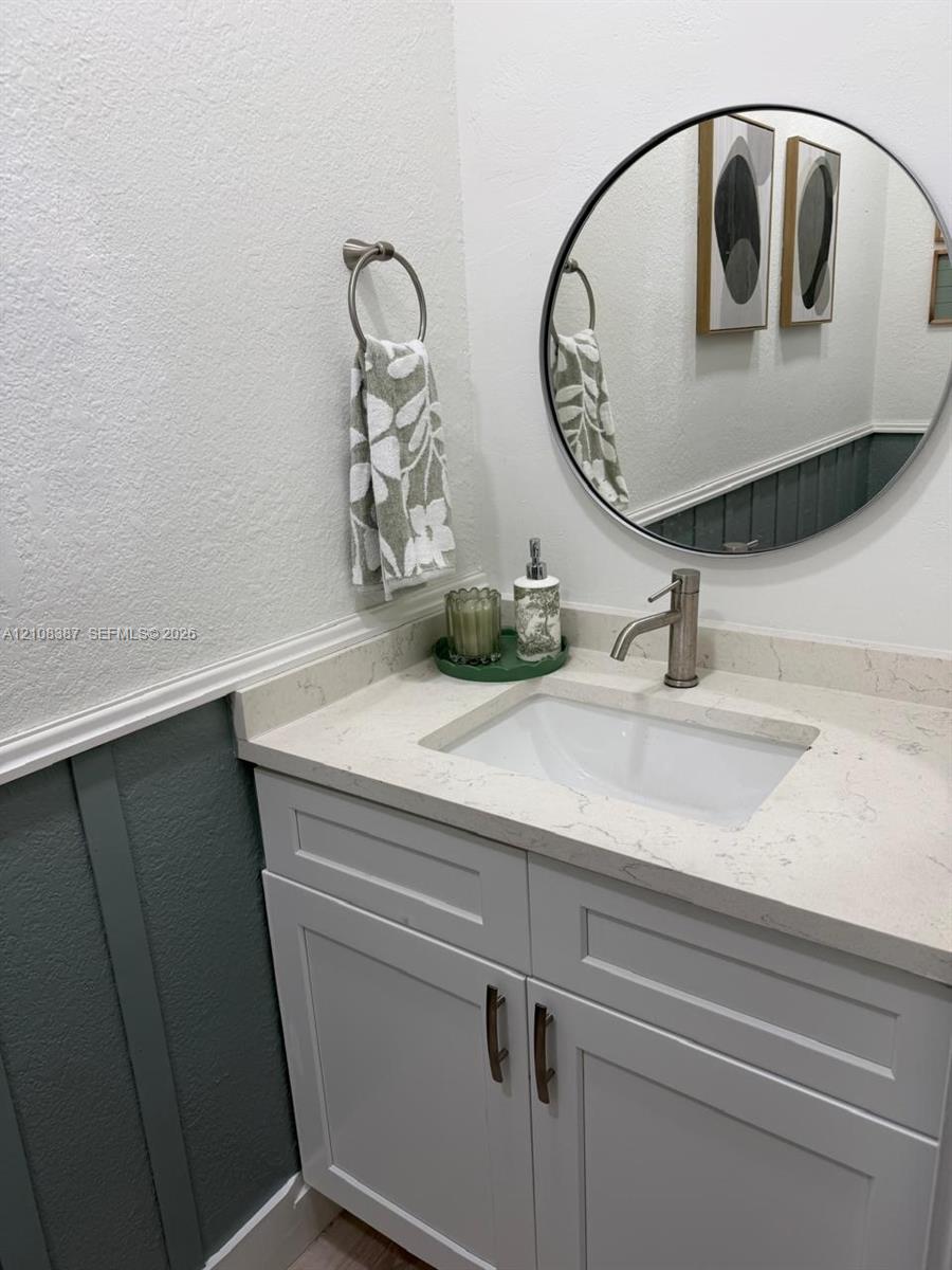 White vanity featuring a quartz countertop, undermount sink, brushed nickel faucet, circular wall-mounted mirror, and decorative wall paneling
