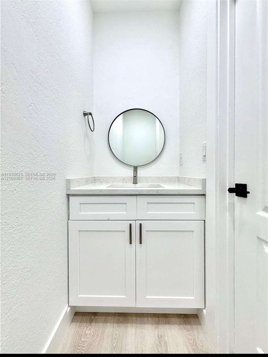 White shaker-style vanity featuring light-toned stone countertop, integrated basin, round wall-mounted mirror, and wood-finish flooring