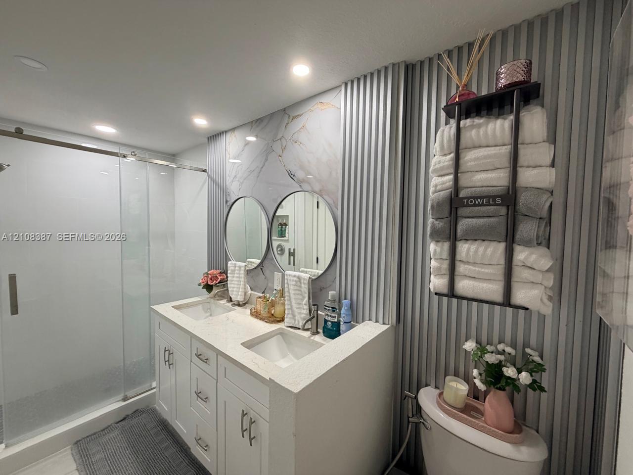 Double vanity featuring a white cabinet base, stone-composite countertop with integrated dual sinks, fluted wall paneling, dual circular mirrors, and a glass-enclosed shower