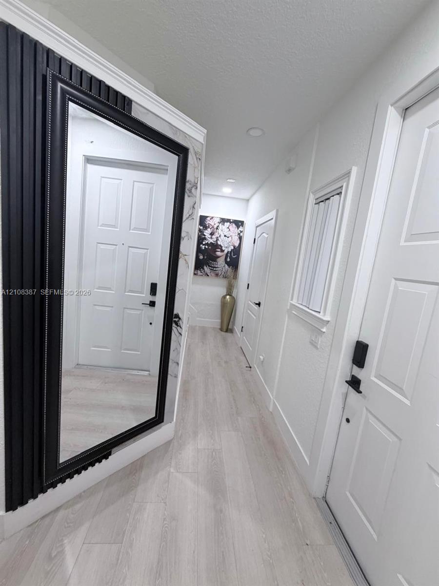 Hallway featuring light-toned wood-finish flooring, decorative wall paneling, textured wall coverings, and multiple paneled interior doors with matte black hardware