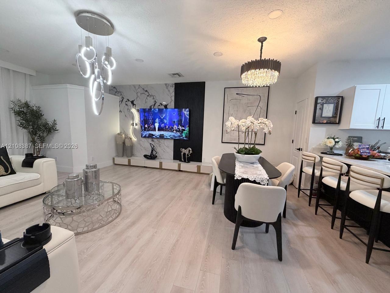Living area featuring light-toned wood-finish flooring, a contemporary multi-ring pendant light, a textured stone-finish accent wall with vertical slat detailing, and a multi-tiered crystal drum chandelier