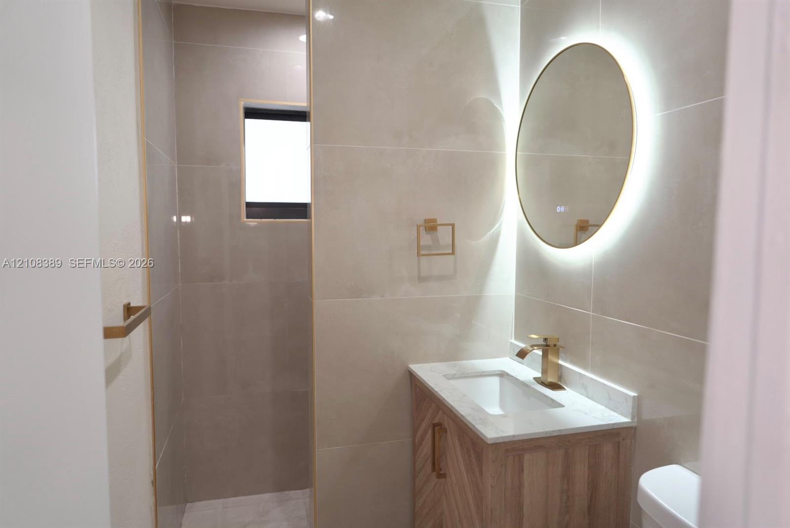 Wood-finish vanity with a stone countertop, wall-mounted circular backlit mirror, brushed gold-tone hardware, and floor-to-ceiling tile work