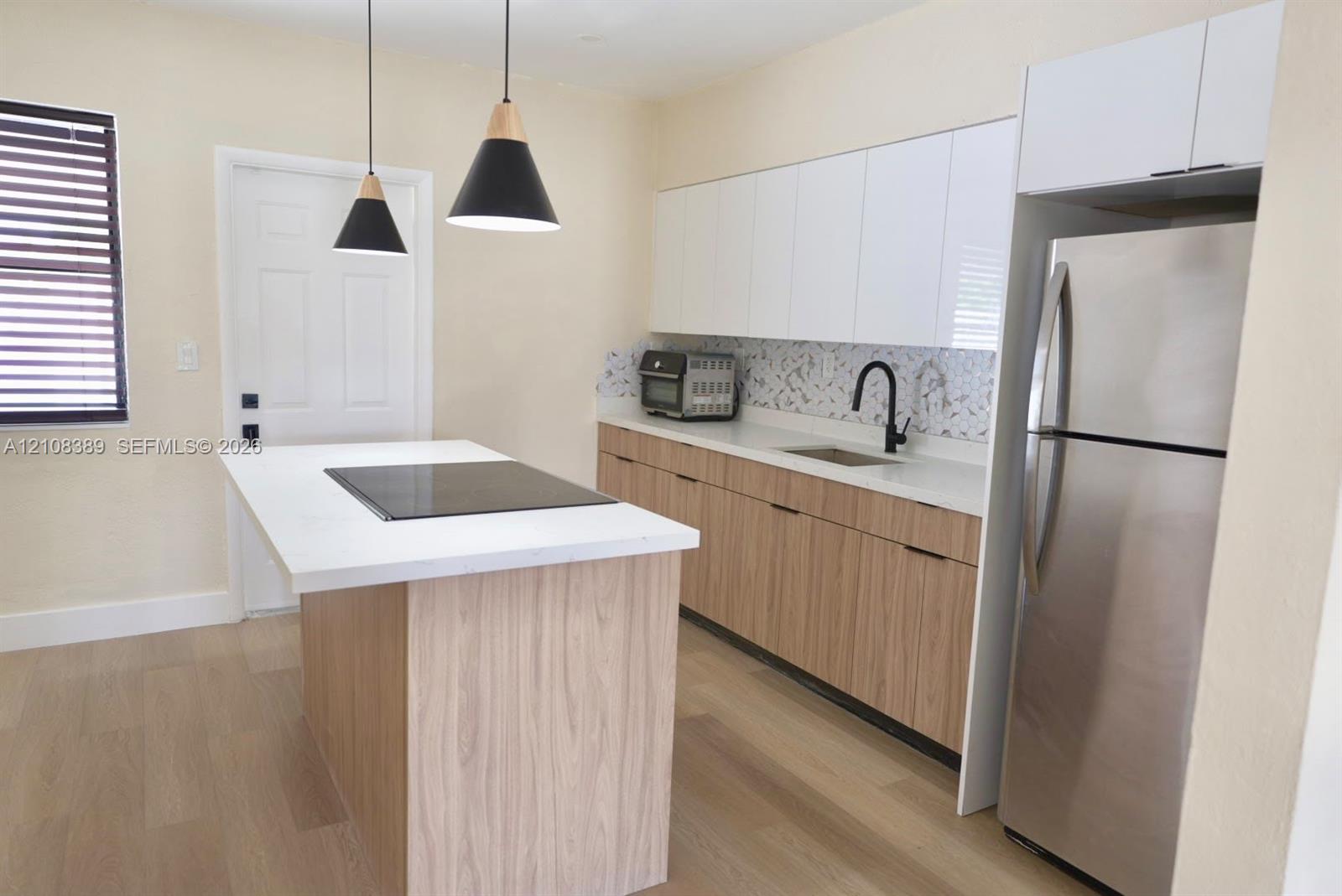 Kitchen featuring a central island with a solid surface countertop and integrated cooktop, wood-finish cabinetry, white high-gloss upper cabinets, patterned backsplash, and matte black pendant lighting