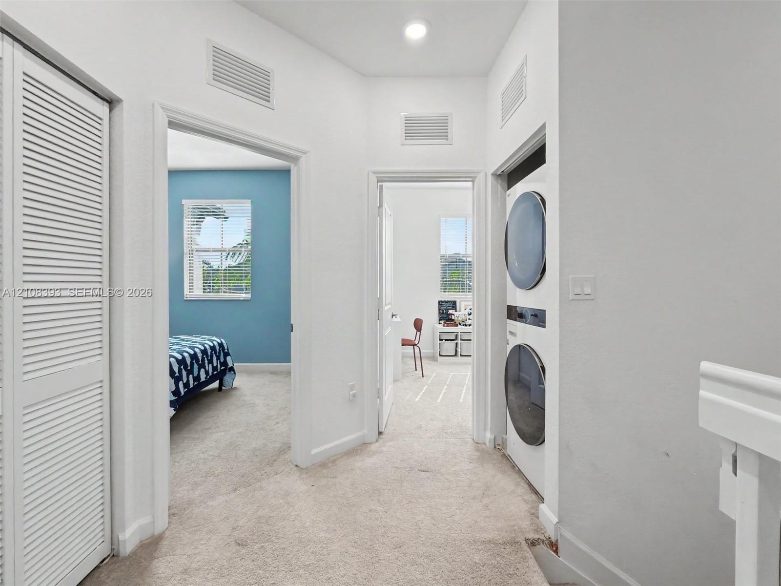 Hallway featuring neutral carpeting, a built-in stacked laundry unit, and louvered closet doors