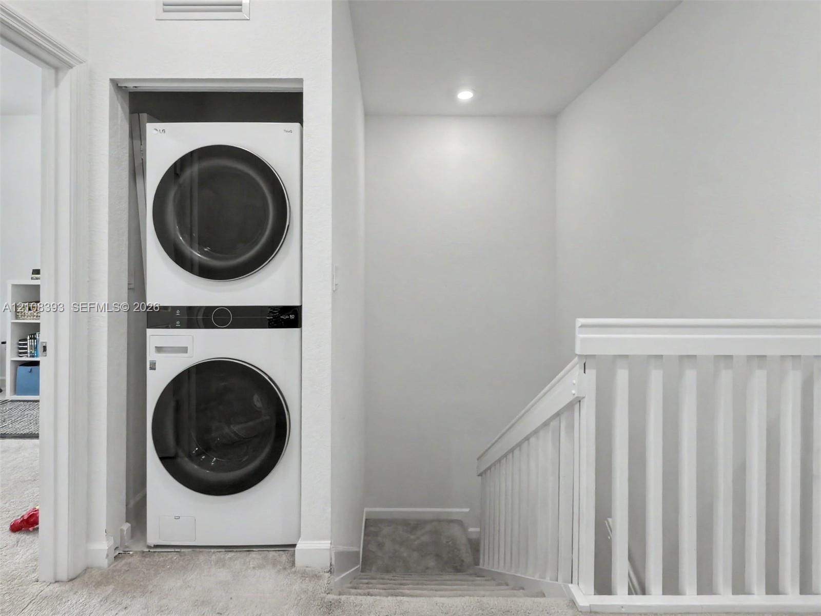 Stacked laundry unit with front-loading appliances, recessed ceiling lighting, and white wooden stair railing