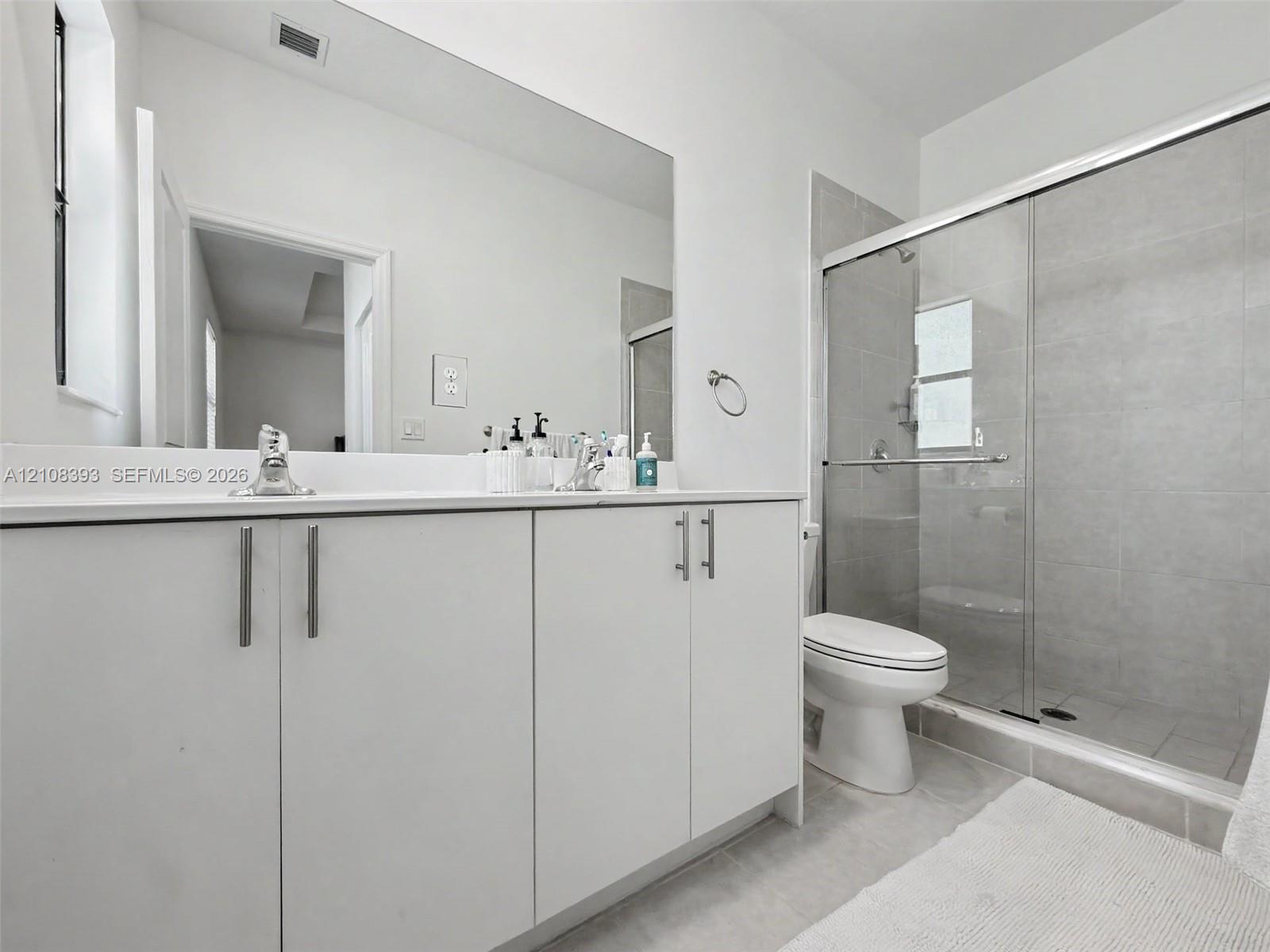 Bathroom featuring white shaker-style cabinetry, solid-surface countertop, frameless glass shower enclosure with neutral-toned wall tile, and wall-to-wall vanity mirror