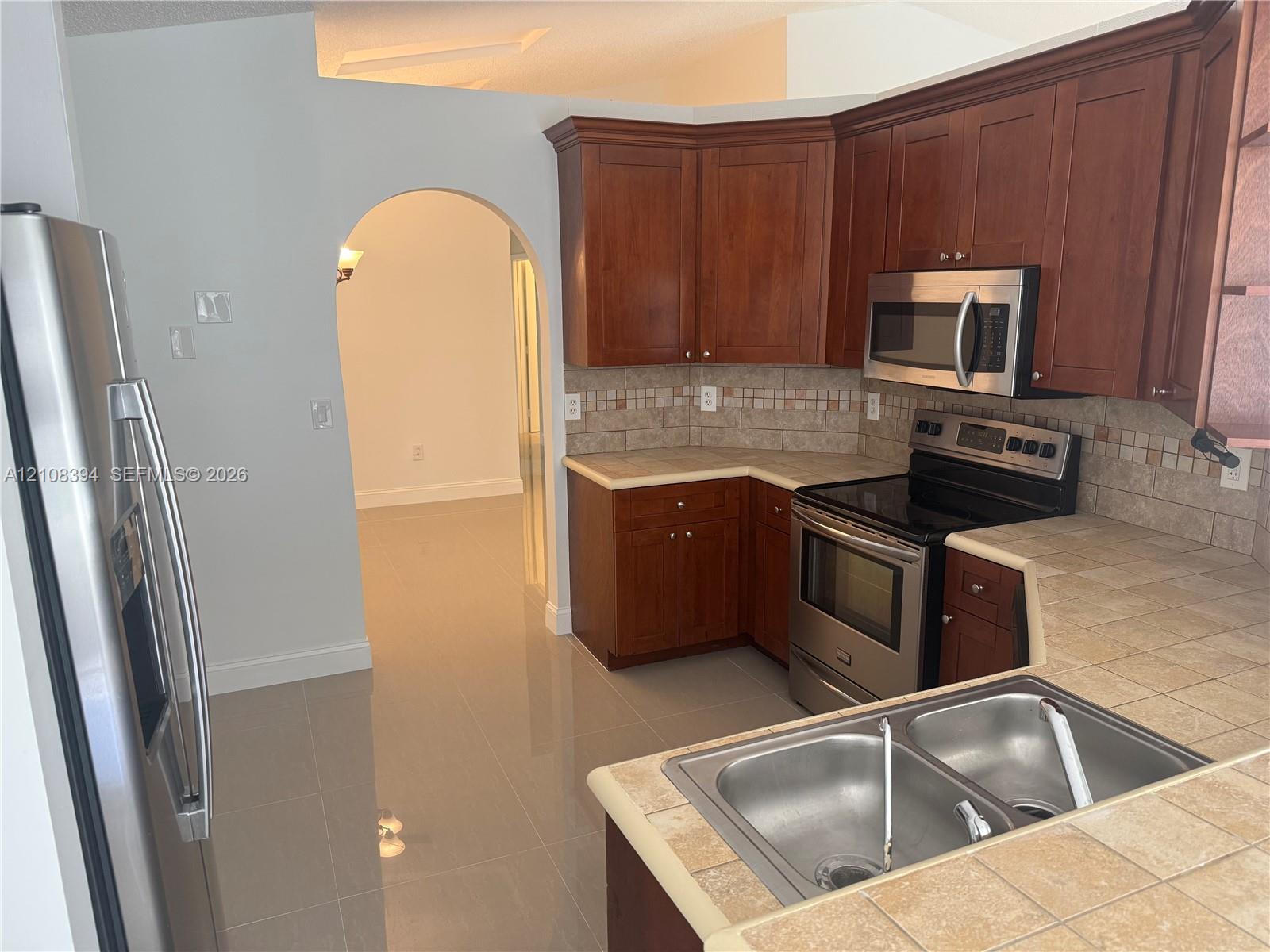 Kitchen featuring cherry-finish cabinetry, stainless steel appliances, tile countertops, a mosaic tile backsplash, and ceramic tile flooring Kitchen featuring cherry-finish cabinetry, stainless steel appliances, tile countertops, a mosaic tile backsplash, and ceramic tile flooring