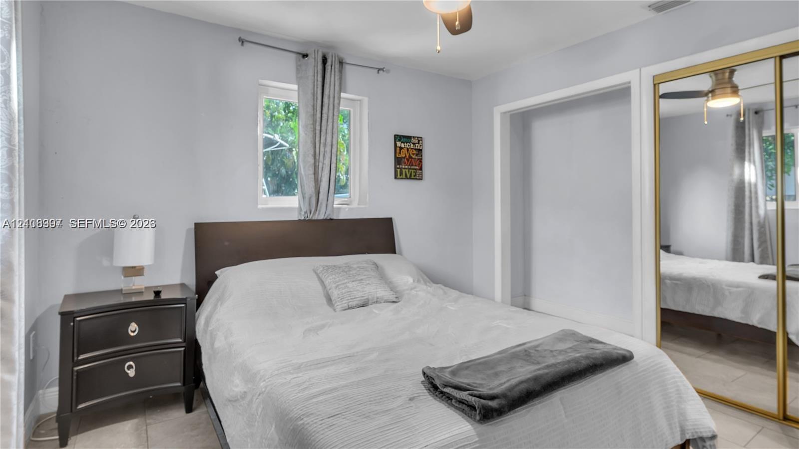Neutral-toned room featuring tiled flooring, a built-in closet with gold-trimmed sliding mirrored doors, a ceiling fan with light fixture, and a window with casing