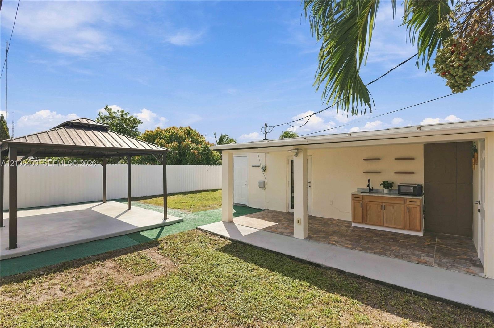 Backyard featuring a detached metal-frame gazebo with a concrete pad, a covered patio with tile flooring, and an outdoor kitchenette with wood-finish cabinetry and a stone-surface countertop Backyard featuring a detached metal-frame gazebo with a concrete pad, a covered patio with tile flooring, and an outdoor kitchenette with wood-finish cabinetry and a stone-surface countertop