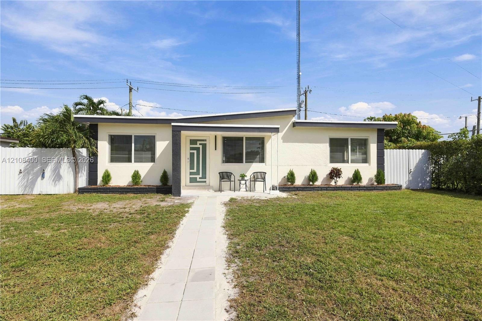 Mid-century modern exterior featuring a low-slope roofline, light-colored stucco facade, geometric-patterned entry door, and white privacy fencing