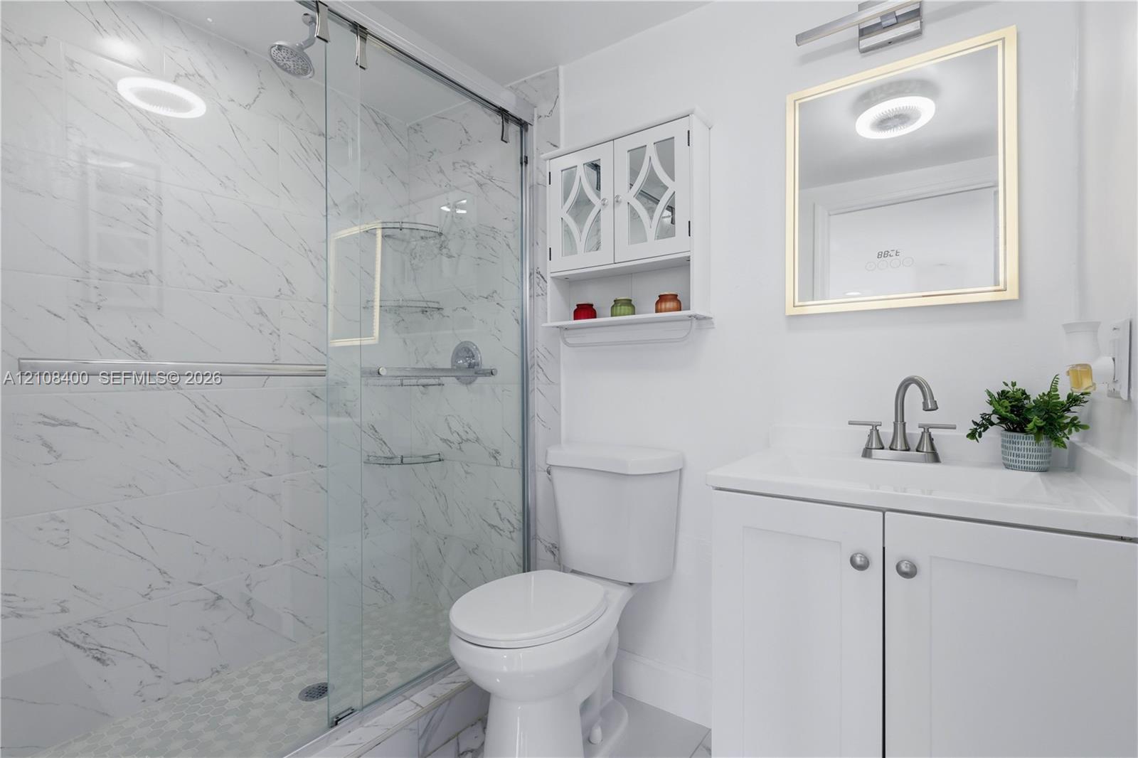 Bathroom featuring a glass-enclosed shower with floor-to-ceiling stone-pattern tile, white vanity cabinet with a integrated sink, polished metal hardware, and wall-mounted storage cabinetry