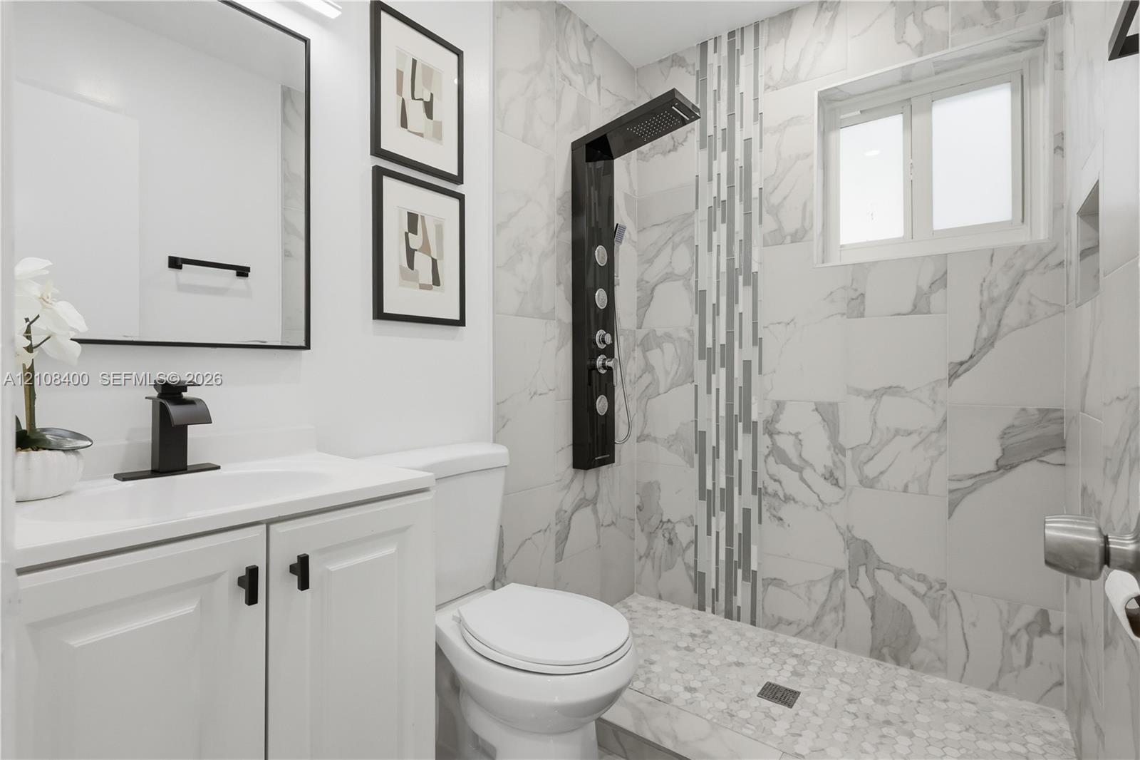 Bathroom featuring large-format wall tile, shower panel system with integrated body jets, walk-in shower with pebble tile flooring, vanity with matte black faucet hardware, and a framed mirror Bathroom featuring large-format wall tile, shower panel system with integrated body jets, walk-in shower with pebble tile flooring, vanity with matte black faucet hardware, and a framed mirror