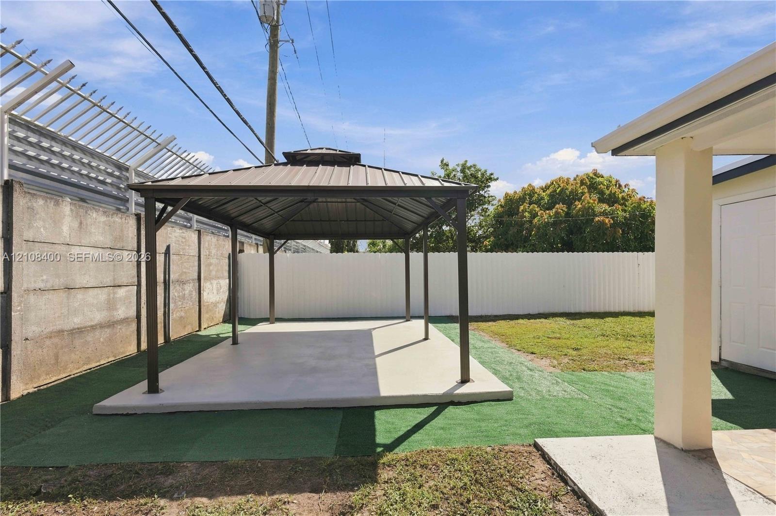 Outdoor area featuring a permanent metal-roofed gazebo, concrete foundation pad, synthetic turf surfacing, and enclosed fencing